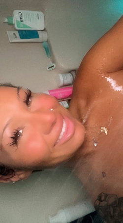 A smiling woman with dark hair and a necklace lies in a bathtub, water dripping from her skin, surrounded by bath