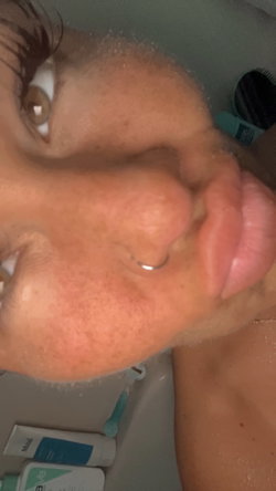 Close-up selfie of a person with wet skin, nose ring, and freckles in a bathroom setting with toiletries visible.