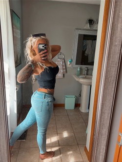 Blonde woman with tattoos takes mirror selfie in bathroom, wearing a black crop top and light blue jeans, barefoot.