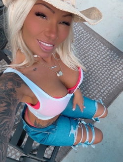 Blonde woman in a white bikini top and ripped jeans sitting outdoors.
