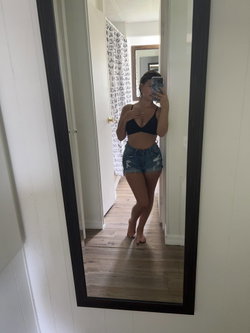 Curvy woman in black bra and distressed denim shorts takes mirror selfie in hallway, barefoot with hand near chest.