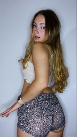A woman with long blonde hair in a white strapless top and patterned shorts poses against a plain background.