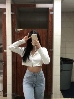 A woman with long dark hair takes a mirror selfie in a bathroom, wearing a white cropped button-up shirt and light blue