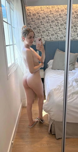 A woman with blonde hair in a bun takes a selfie in a bedroom with patterned wallpaper.