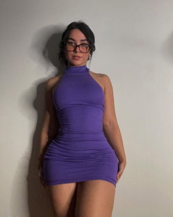 Curvy woman with dark hair and glasses wears a tight purple halter dress, leaning against a plain wall.