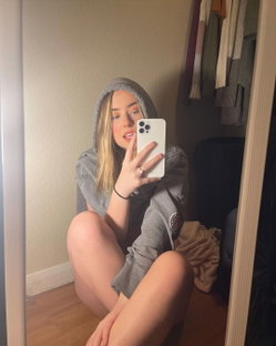 Blonde woman in a gray hoodie sits on the floor, taking a mirror selfie with her phone, legs bent and bare.