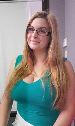 A woman with long blonde hair and glasses wears a teal top, smiling softly in an indoor setting.