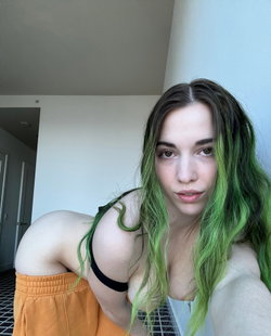 Person with green-tipped hair in a black bra and orange shorts leans on a surface.