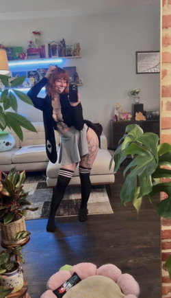 Redhead in glasses and open black cardigan poses for a mirror selfie in her living room.