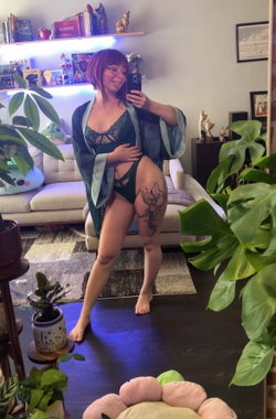 Curvy woman with short red hair and glasses poses in green lingerie and robe in a living room.