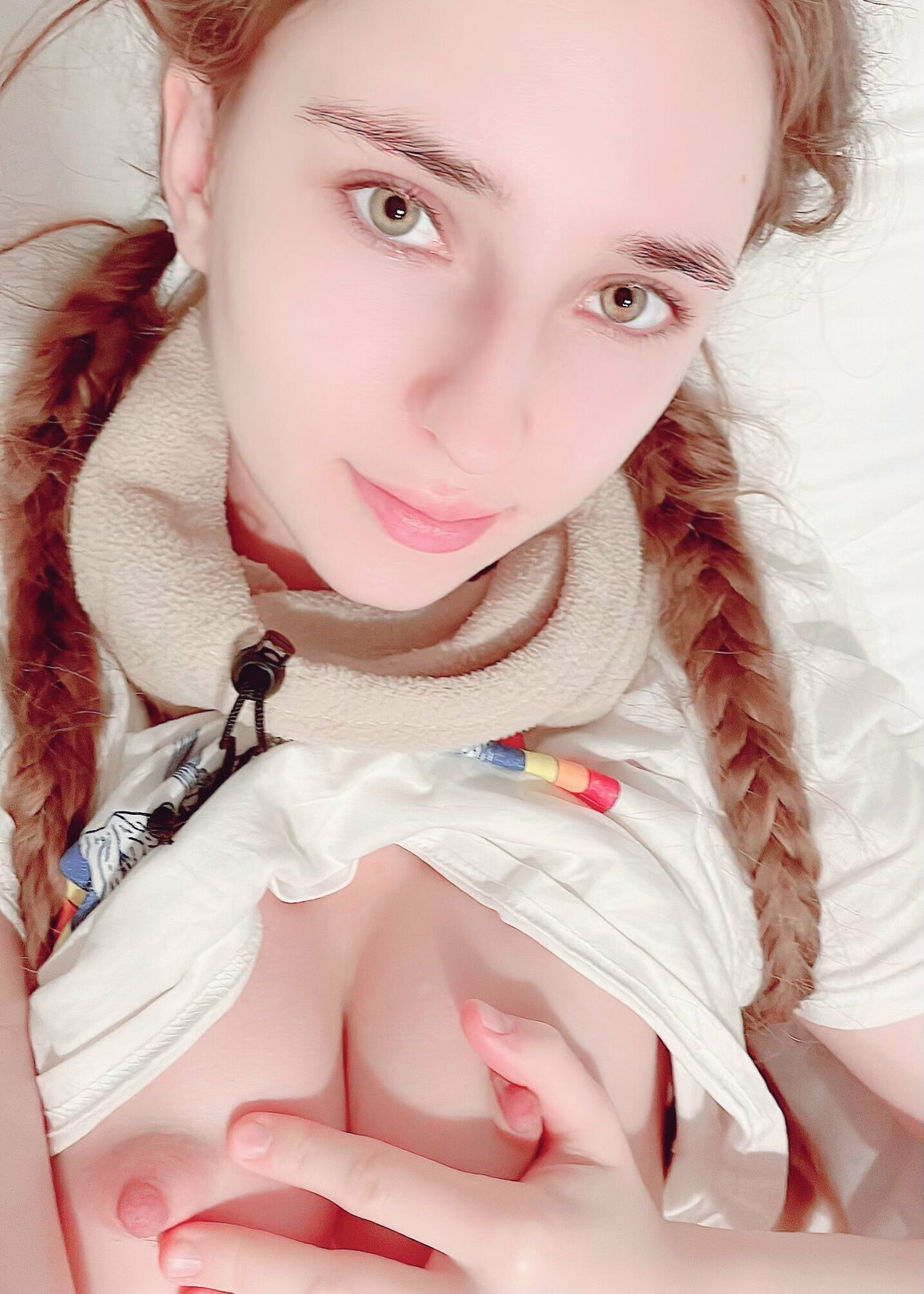 Woman with braided auburn hair in white fleece taking a selfie on bed, top pulled down.