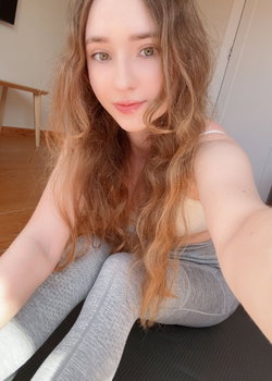 Woman with long wavy brown hair in a beige crop top and gray leggings taking a selfie indoors.