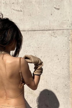 Back view of a slim woman in beige lingerie and gloves posing against a concrete wall.