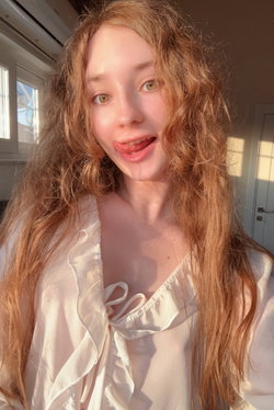 Redhead with long wavy hair wearing a white ruffled blouse taking a selfie indoors.
