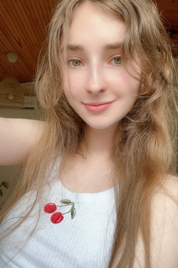 Blonde woman with long hair takes a selfie indoors in a white tank top with cherry embroidery.