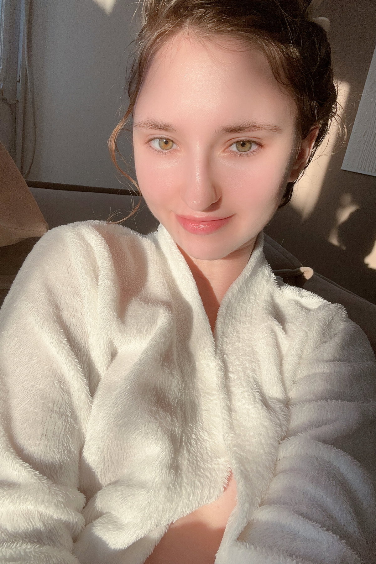 A young woman with brown hair and green eyes smiles softly while wearing a white fluffy robe, sitting indoors in