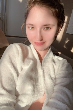 A young woman with brown hair and green eyes smiles softly while wearing a white fluffy robe, sitting indoors in