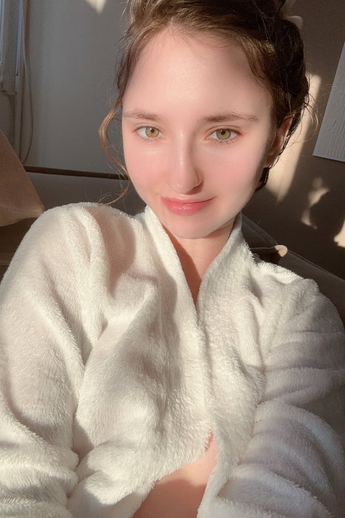 A young woman with brown hair and green eyes smiles softly while wearing a white fluffy robe, sitting indoors in