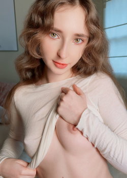 A young woman with wavy brown hair and a white sweater poses in a cozy setting.