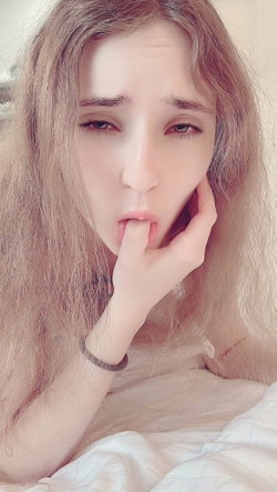 Long-haired blonde woman in a white top lies in bed taking a selfie.