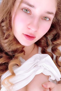 Woman with long wavy brown hair and green eyes takes a close-up selfie in a white top.