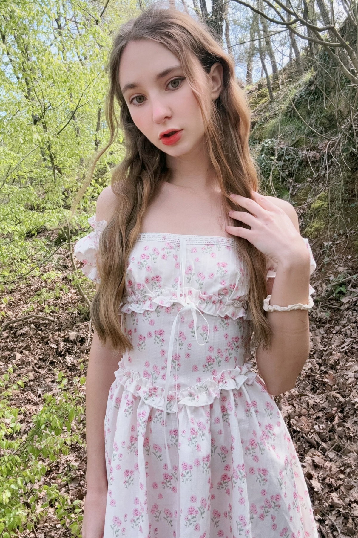 Young woman with flowing brown hair in a white floral dress poses outdoors in the woods.