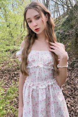 Young woman with flowing brown hair in a white floral dress poses outdoors in the woods.