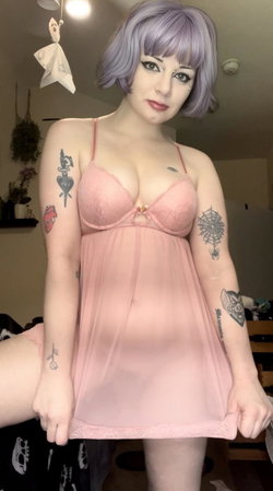 Woman with lavender bob wig and multiple arm tattoos wears a pink lace bra and sheer dress, posing indoors.