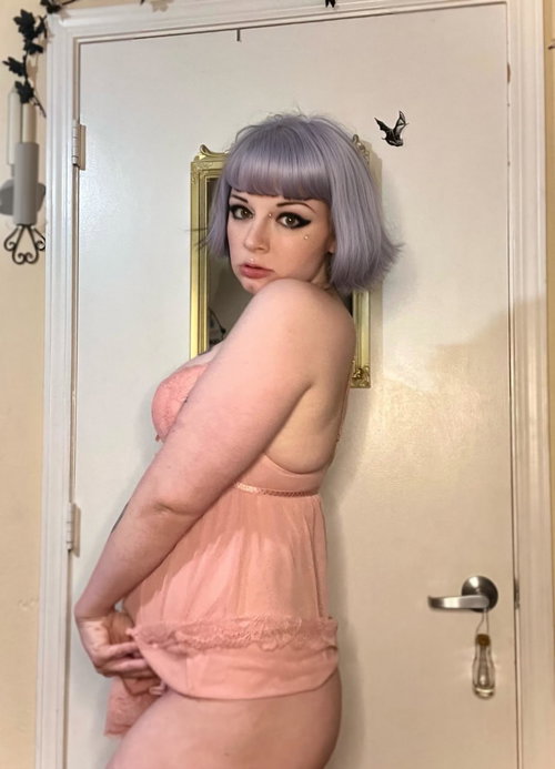 A woman with short lavender hair and dark makeup poses in a peach lace top, showing her back and side against a white