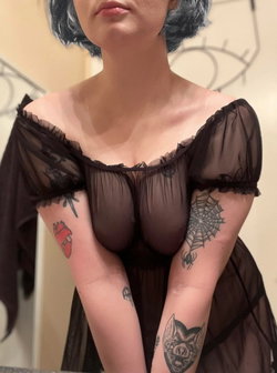 Woman with short dark hair wearing sheer black off-shoulder top, showing tattoos on arms, indoors.