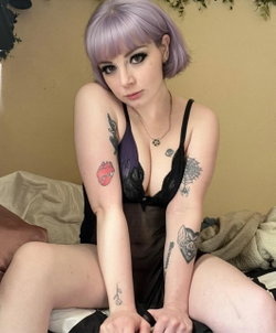 Woman with lavender bob hair and multiple arm tattoos wears black lace lingerie while sitting on a bed with soft
