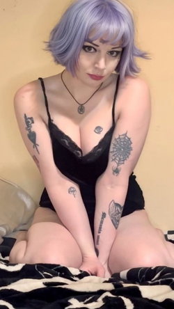 Woman with purple bob wig, multiple arm tattoos, wearing black lace lingerie, sitting on a patterned blanket indoors.