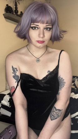Woman with purple bob wig, multiple arm tattoos, wearing black lace-trimmed velvet top, sitting on bed with striped