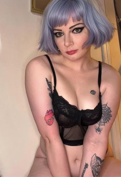 Woman with short purple hair, wearing black lace lingerie, multiple arm tattoos, posing indoors.