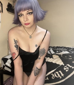 Woman with purple bob wig, multiple arm tattoos, wearing black lace top, sitting on bed with patterned bedding.