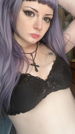 Woman with purple hair and dark makeup wears a black lace bra, showing a tattoo on her chest and arm, in an indoor