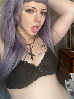 Woman with purple hair and dark makeup wears a black lace bra, sticks out her tongue, and poses in a bedroom.