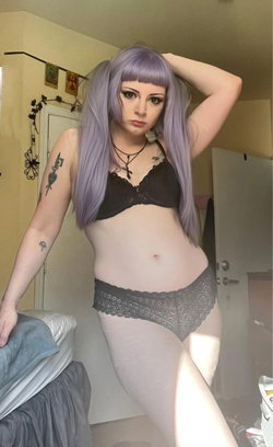 Person with purple wig and dark lace lingerie poses in a bedroom, arm raised, tattoos visible on arm.