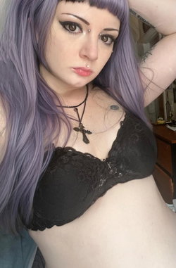 Woman with purple hair and black bra poses in a bedroom.