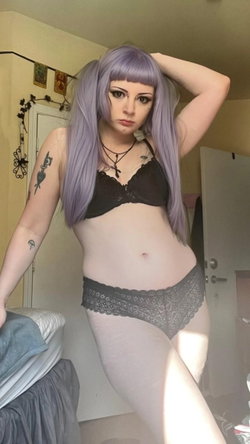 Person with purple wig and dark makeup wears black lace bra and gray lace underwear, posing in a bedroom with arm