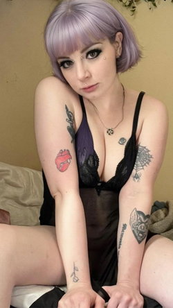 Woman with lavender bob hair and multiple arm tattoos wears black lace lingerie, sitting on a bed with a neutral