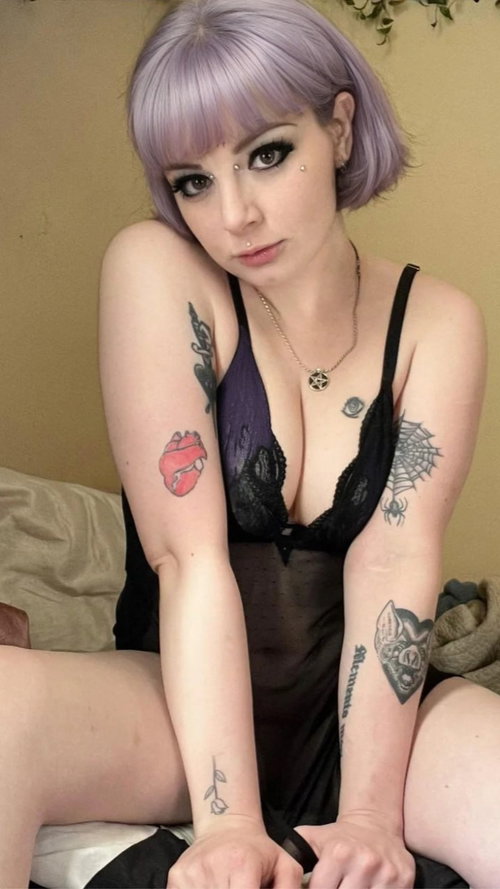 Woman with lavender bob hair and multiple arm tattoos wears black lace lingerie, sitting on a bed with a neutral