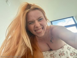 Redheaded woman with long hair, smiling playfully in a floral top, taking a selfie indoors near a window.
