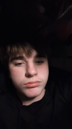 Slim guy with shaggy brown hair in black hoodie, dim close-up selfie.