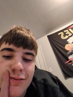 Slim person with bangs wearing black hoodie gestures in bedroom with wall banner.