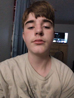 Close-up selfie of man with tousled brown hair wearing beige v-neck t-shirt in bedroom.