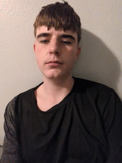 Man with short brown bangs wearing black scoop-neck shirt, neutral side gaze against plain wall.