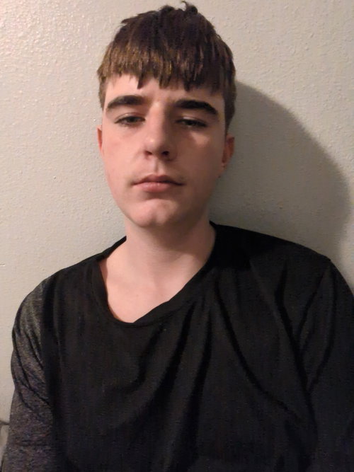 Man with short brown bangs wearing black scoop-neck shirt, neutral side gaze against plain wall.