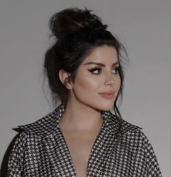 Brunette woman with dark hair in a messy bun, wearing a black and white houndstooth blouse, posing against a plain gray