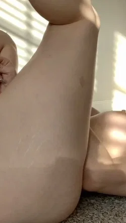 Slim legs raised in a sunlit room with a hand resting on the thigh and thin string visible.